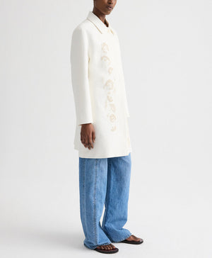 A person wears the TWP Edinburgh Coat with light floral embroidery, paired with blue wide-leg jeans and brown sandals, standing against a plain white background.