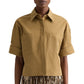 A person wearing the TWP My Newest Mistake Shirt, a boxy fit tan short-sleeve Italian cotton button-up with cuffed sleeves and two chest pockets, paired with a brown and gold pleated skirt.