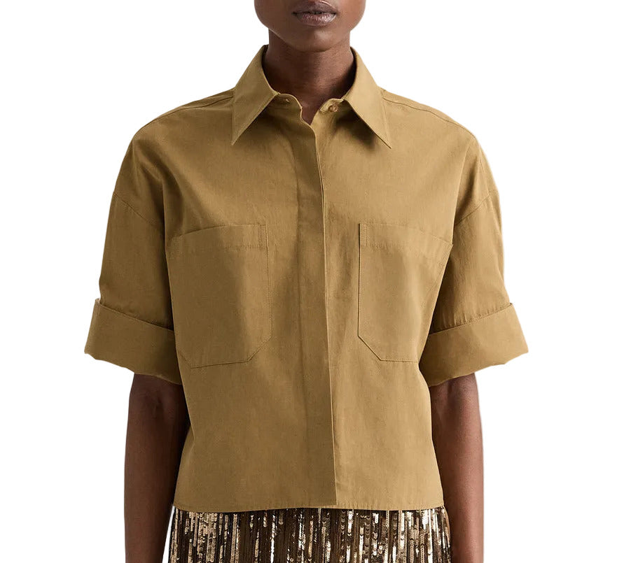A person wearing the TWP My Newest Mistake Shirt, a boxy fit tan short-sleeve Italian cotton button-up with cuffed sleeves and two chest pockets, paired with a brown and gold pleated skirt.