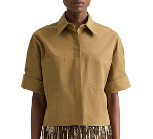 A person wearing the TWP My Newest Mistake Shirt, a boxy fit tan short-sleeve Italian cotton button-up with cuffed sleeves and two chest pockets, paired with a brown and gold pleated skirt.
