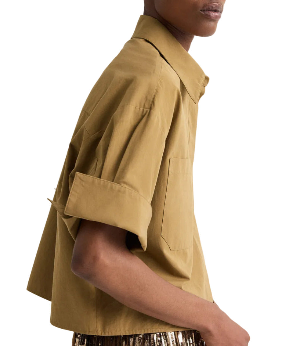 A person is shown from the side wearing TWP's My Newest Mistake Shirt, a tan, loose-fitting, boxy short-sleeve button-up with rolled sleeves and a collar, set against a plain background.