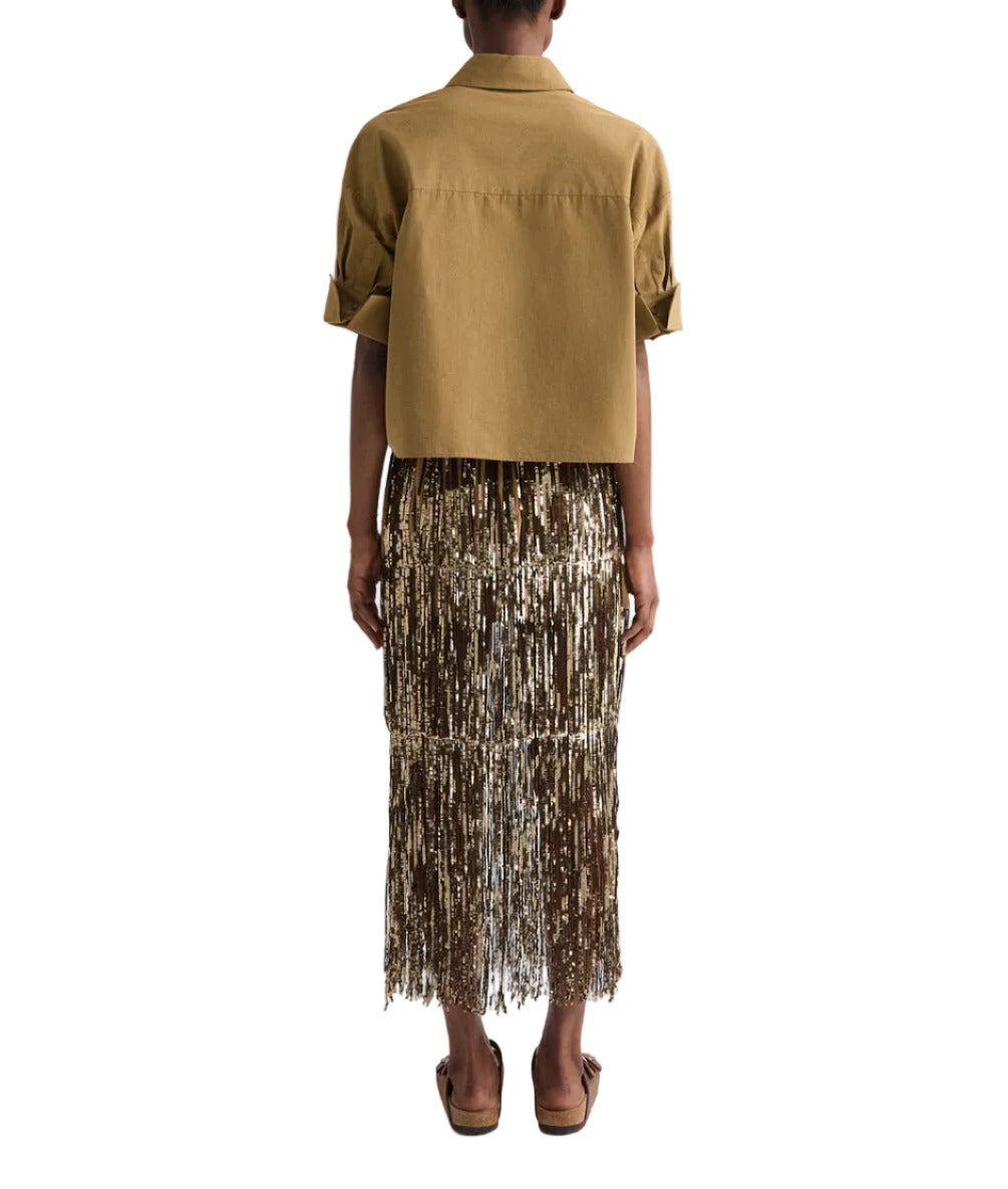 A person stands with their back to the camera against a white background, wearing the TWP My Newest Mistake Shirt—a short-sleeve boxy button-up—layered under a cropped tan jacket, paired with a sheer brown fringe skirt and brown sandals.