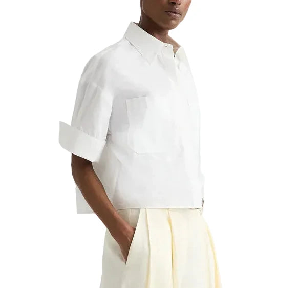 A person wearing the TWP My Newest Mistake Shirt—relaxed fit, short-sleeve, button-up Italian cotton with a collar—paired with cream pleated trousers, stands with one hand in a pocket against a plain background.