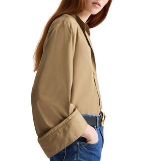 A person with long red hair models the TWP Beau Shirt in tan Supima cotton, styled with rolled sleeves and blue jeans, shown from the side against a white background.