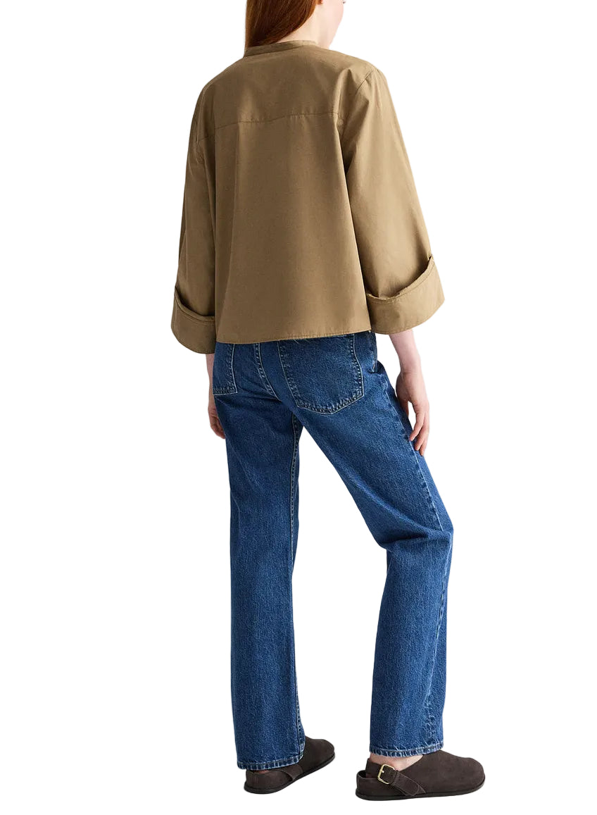 A person stands with their back to the camera, wearing a tan jacket over the TWP Beau Shirt, blue jeans, and brown shoes.