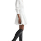 A person wearing the TWP Mila Dress, a long-sleeve white cotton shirt dress, with knee-high black boots stands against a white background.