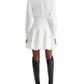 A person stands facing away in the TWP Mila Dress—a white cotton shirt dress with a flared skirt and corset waist—paired with black knee-high boots, set against a white background.