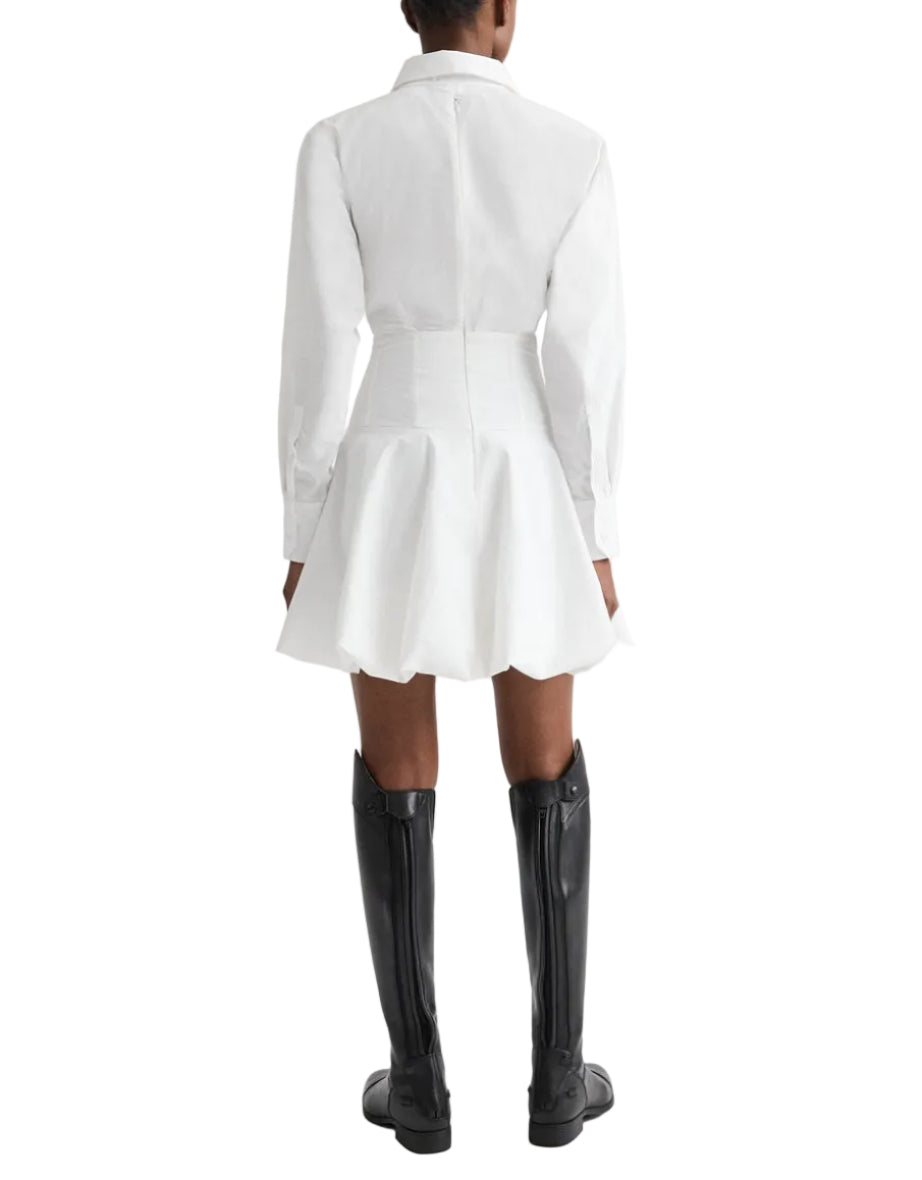 A person stands facing away in the TWP Mila Dress—a white cotton shirt dress with a flared skirt and corset waist—paired with black knee-high boots, set against a white background.