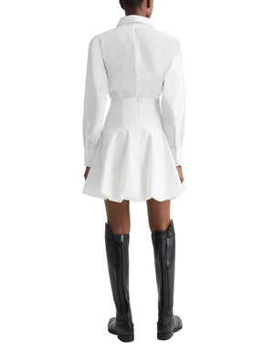 A person stands facing away in the TWP Mila Dress—a white cotton shirt dress with a flared skirt and corset waist—paired with black knee-high boots, set against a white background.