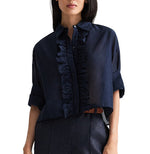 A woman wearing the TWP Next Ex Ruffle Top, a navy blue short-sleeve button-down blouse featuring ruffle detailing down the front and rolled-up sleeves, paired with dark high-waisted pants.