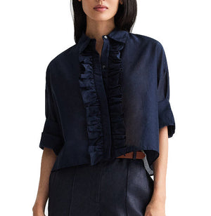 A woman wearing the TWP Next Ex Ruffle Top, a navy blue short-sleeve button-down blouse featuring ruffle detailing down the front and rolled-up sleeves, paired with dark high-waisted pants.