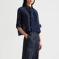 A woman stands with her hands in her pockets against a plain background, wearing the TWP Next Ex Ruffle Top—a navy cotton-silk organza short-sleeve button-down blouse with ruffled detailing—paired with matching navy high-waisted pants.