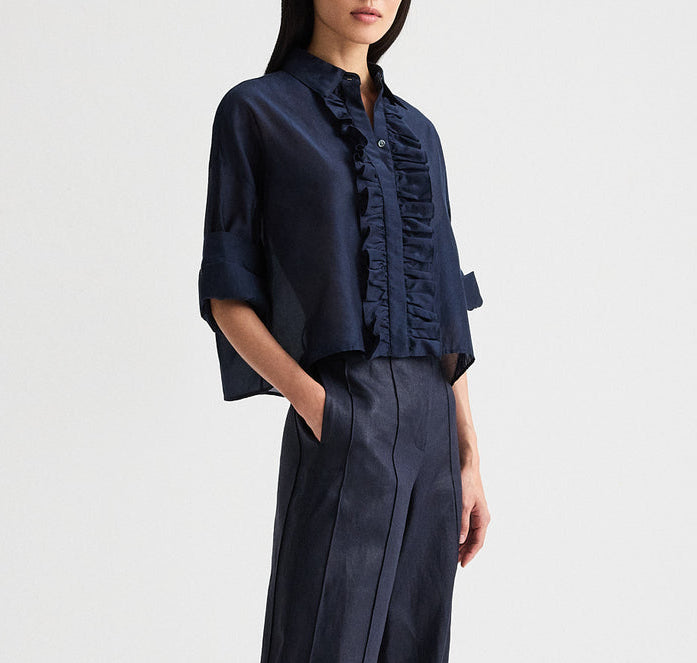 A woman stands with her hands in her pockets against a plain background, wearing the TWP Next Ex Ruffle Top—a navy cotton-silk organza short-sleeve button-down blouse with ruffled detailing—paired with matching navy high-waisted pants.