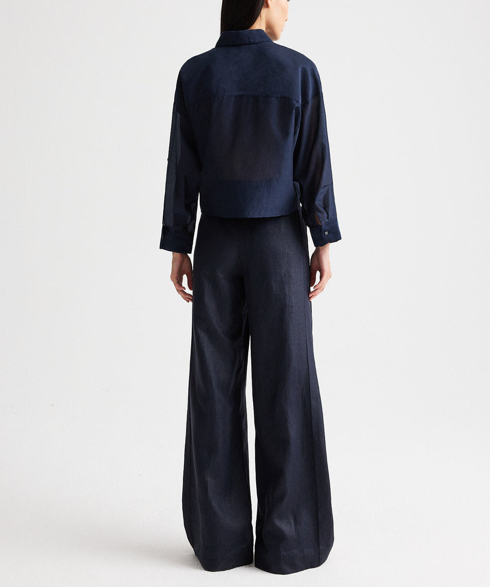A person stands with their back to the camera, wearing the TWP Next Ex Ruffle Top—a loose-fitting, dark blue long-sleeve top—paired with matching wide-leg pants featuring a cropped boxy silhouette, set against a plain light background.