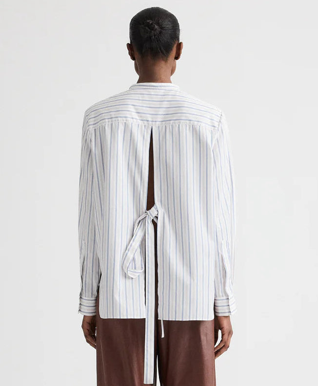Person facing away, wearing the TWP Tied Down Stripe Shirt—white with blue vertical stripes, back slit and tie detail—paired with brown pants.