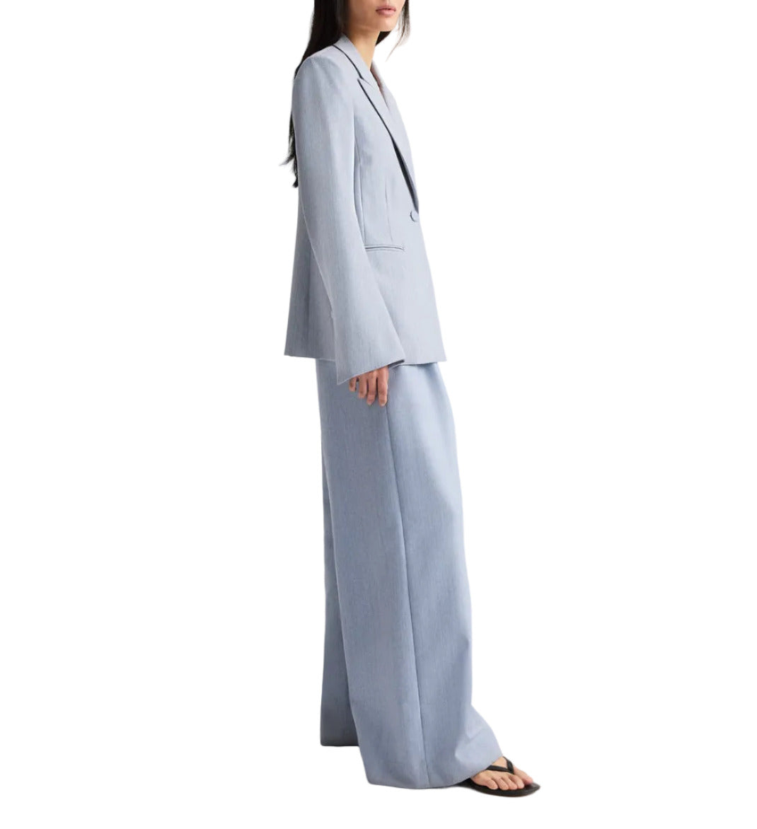 A woman wears the TWP Her Fit Blazer in light blue, paired with wide-leg trousers and black sandals, standing against a white background.