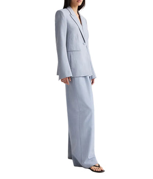 A woman stands against a white background, wearing the TWP Avery Pant—light blue, oversized wool suiting with wide-leg pleated trousers—and black open-toe sandals.