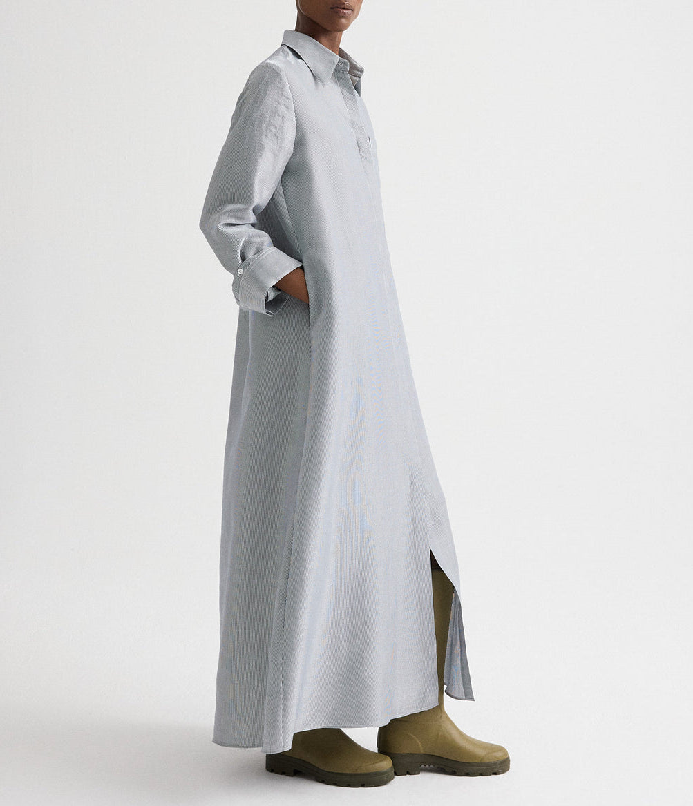 A person wears the TWP Jenny's Glossy Mini Stripe Gown, a long, light gray collared oversized shirtdress with side pockets and front slit, styled with olive green boots against a plain white background.