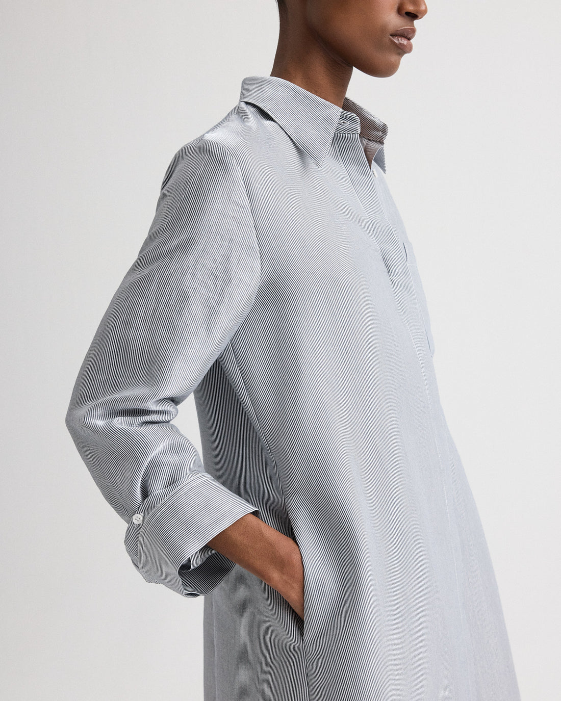 Model wears TWP’s Jenny's Glossy Mini Stripe Gown—a light grey, long-sleeved, collared shirting dress with side pockets—photographed from the shoulders down against a plain background.