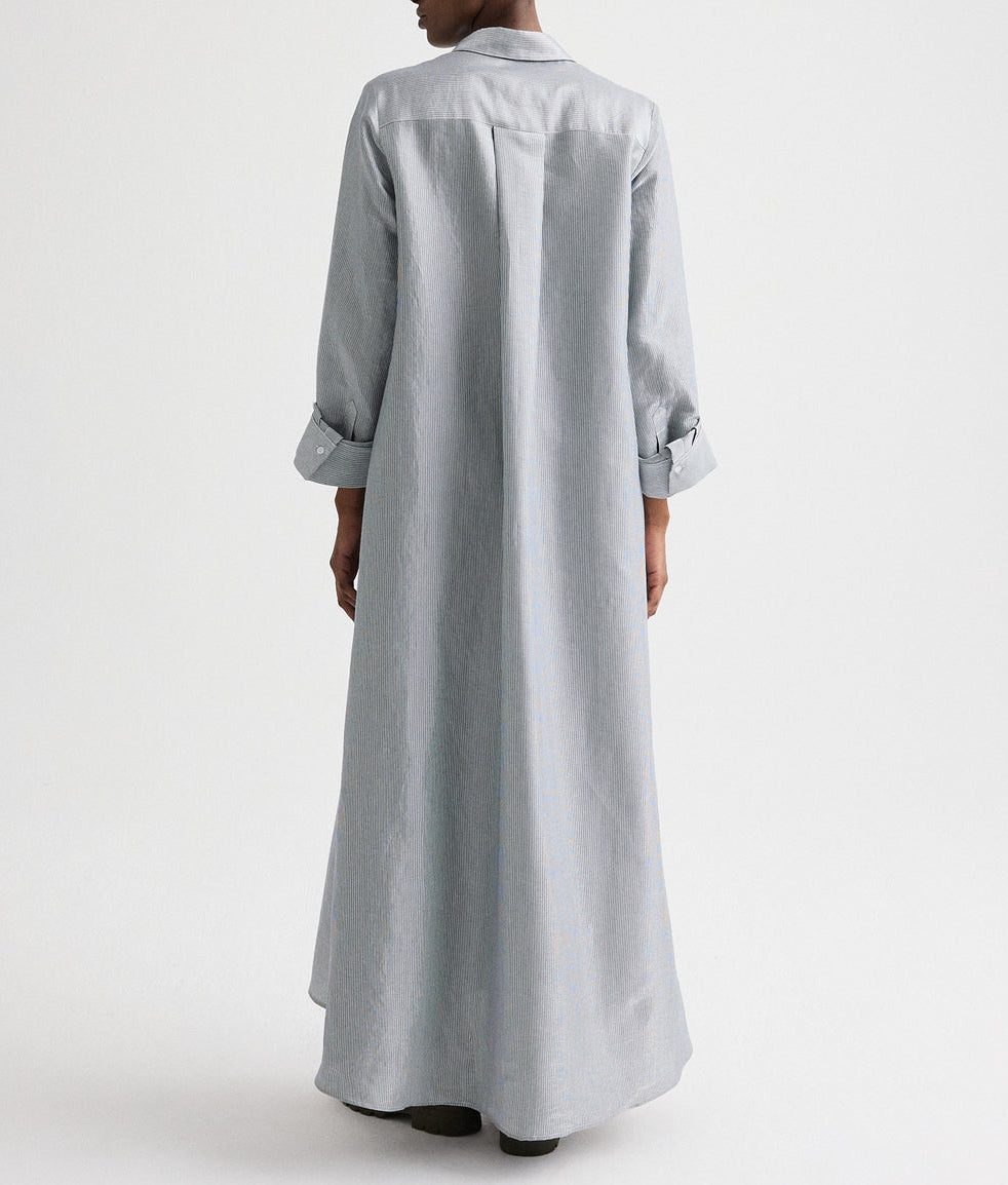 A person stands with their back to the camera in TWP’s Jenny's Glossy Mini Stripe Gown, an oversized light gray cotton shirtdress with long sleeves and subtle glossy mini stripes, shown against a plain white background.