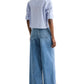 Person standing with back to camera, wearing the TWP Next Ex Stripe Shirt (light blue striped, cropped boxy fit), wide-leg blue jeans, and brown shoes.