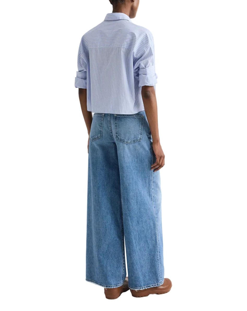 Person standing with back to camera, wearing the TWP Next Ex Stripe Shirt (light blue striped, cropped boxy fit), wide-leg blue jeans, and brown shoes.