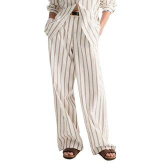 A person wears TWP's Hudson St. Cotton Stripes Pant—a white, wide-leg, brown pinstripe style—paired with a matching button-up shirt, brown sandals, and a black belt. Hands are casually tucked in pockets.