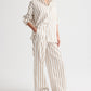 Model in a loose-fitting white shirt and TWP Hudson St. Cotton Stripes Pant—wide-leg, mid-rise pants with tan vertical stripes—paired with brown sandals, posing against a plain white background.