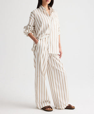 Model in a loose-fitting white shirt and TWP Hudson St. Cotton Stripes Pant—wide-leg, mid-rise pants with tan vertical stripes—paired with brown sandals, posing against a plain white background.