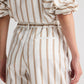 Model wearing the Hudson St. Cotton Stripes Pant by TWP, a wide-leg, mid-rise style with vertical beige stripes, paired with a matching white pinstripe shirt. Sleeves are rolled up; shown from back and side views.