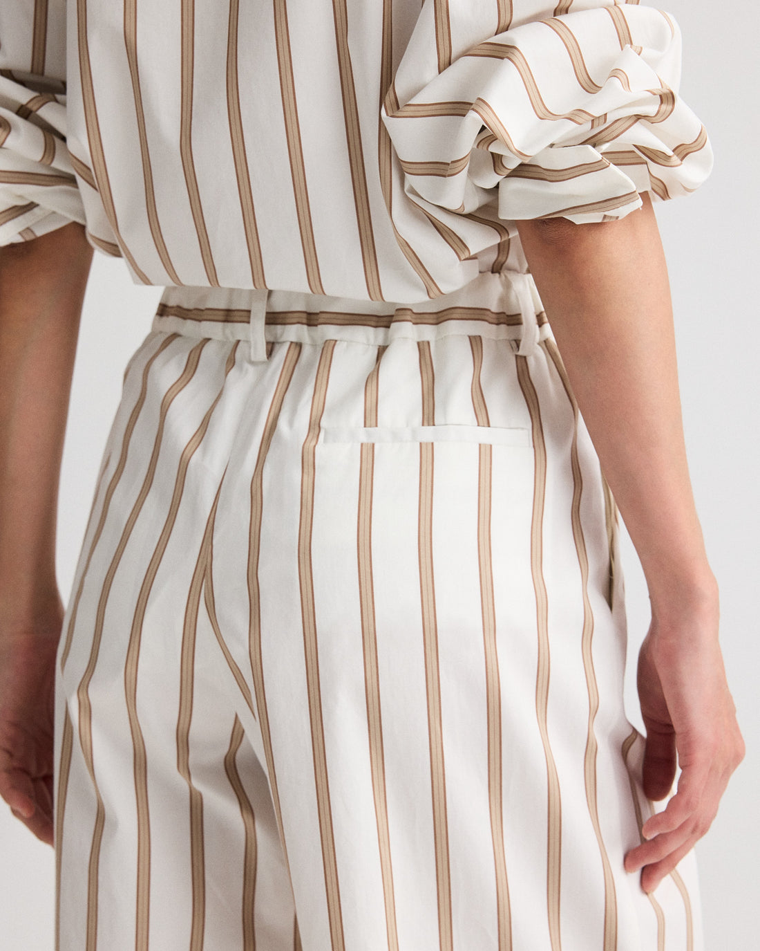 Model wearing the Hudson St. Cotton Stripes Pant by TWP, a wide-leg, mid-rise style with vertical beige stripes, paired with a matching white pinstripe shirt. Sleeves are rolled up; shown from back and side views.