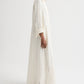 A person wearing TWP's Jenny's Ladder Weave Linen Gown—a long, white robe with vertical perforated stripes and rolled-up sleeves—pairs it with brown sandals against a plain background, highlighting its relaxed silhouette.