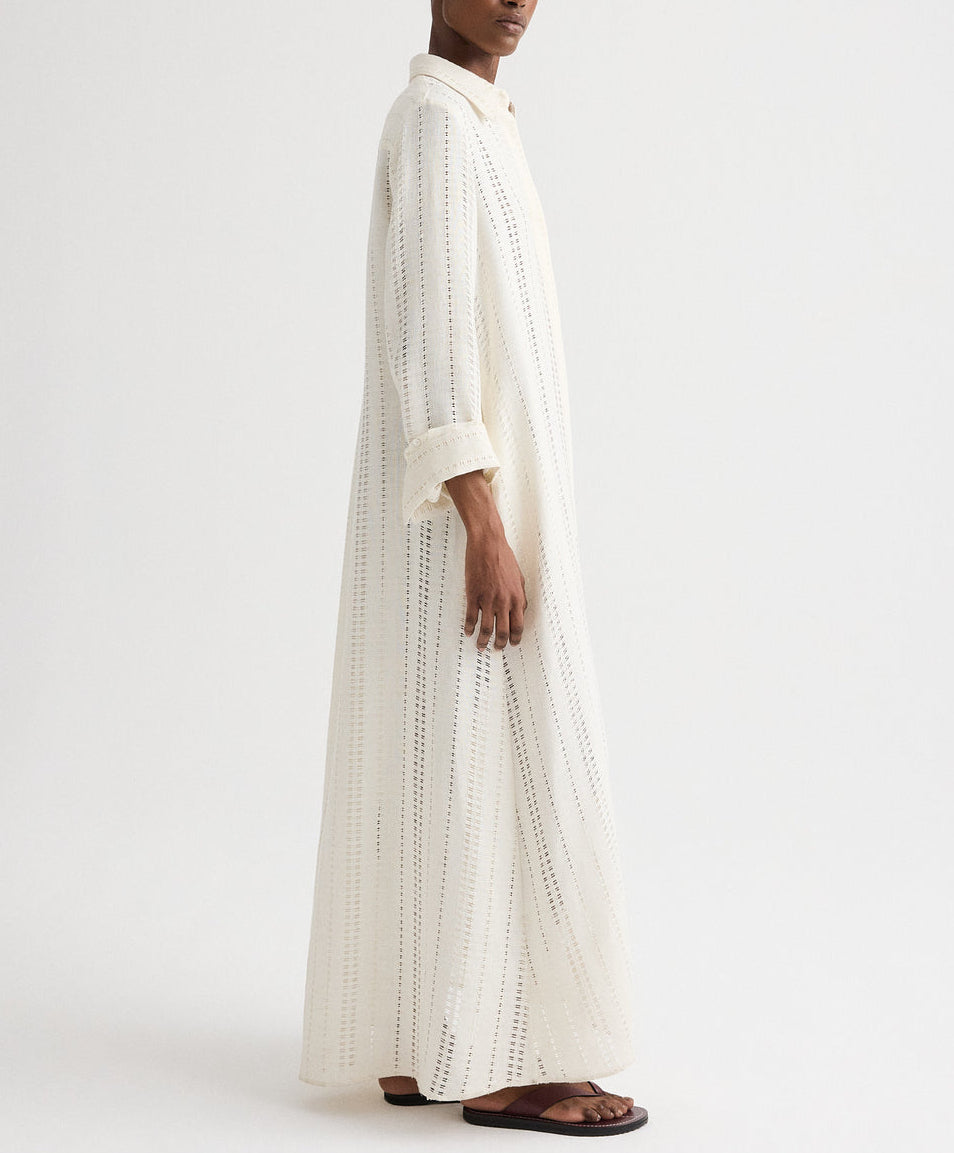 A person wearing TWP's Jenny's Ladder Weave Linen Gown—a long, white robe with vertical perforated stripes and rolled-up sleeves—pairs it with brown sandals against a plain background, highlighting its relaxed silhouette.