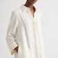 Someone wears TWP's Jenny's Ladder Weave Linen Gown, a cream-colored, oversized maxi shirtdress with subtle vertical cutout stripes and a relaxed silhouette.