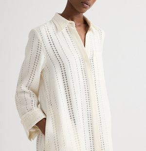 Someone wears TWP's Jenny's Ladder Weave Linen Gown, a cream-colored, oversized maxi shirtdress with subtle vertical cutout stripes and a relaxed silhouette.