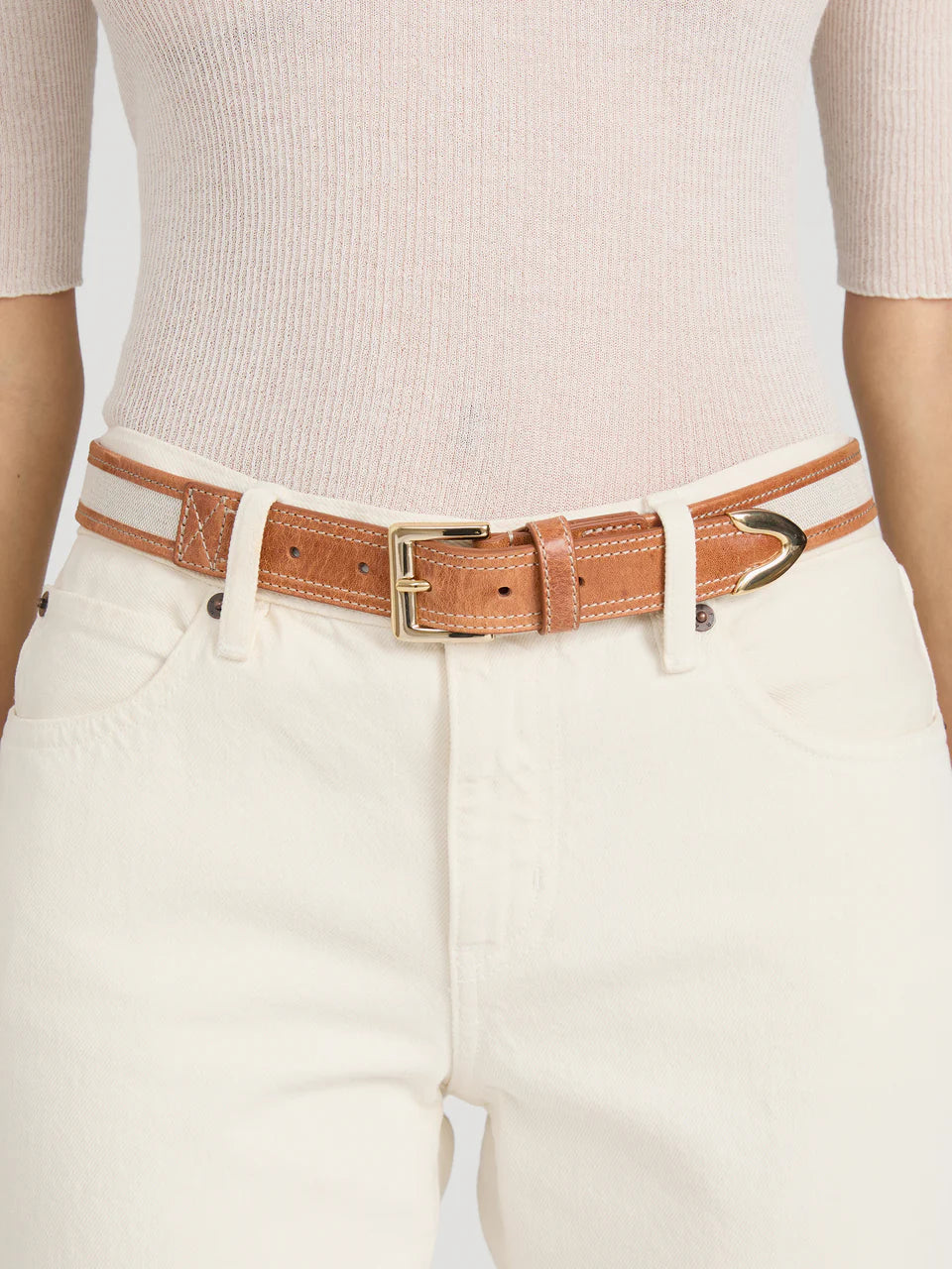 Person wearing high-waisted white jeans with Frame's The Riviera Belt, a slim cow leather belt with a polished buckle, over a fitted light beige ribbed top.
