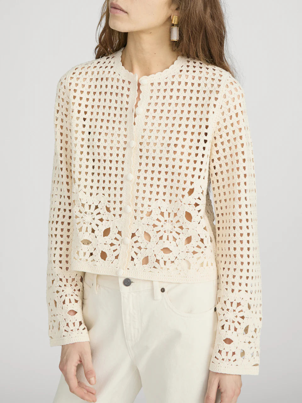 A woman wearing Frame’s The Postcard Cardi, a cream-colored, long-sleeve crochet cardigan with a round neckline, paired with cream pants.