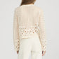 A woman models The Postcard Cardi by Frame—a cream, long-sleeve crochet cardigan with geometric and floral designs—paired with white pants, shown from the back against a plain background.