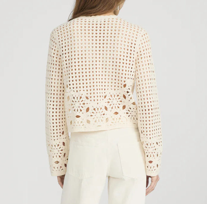 A woman models The Postcard Cardi by Frame—a cream, long-sleeve crochet cardigan with geometric and floral designs—paired with white pants, shown from the back against a plain background.