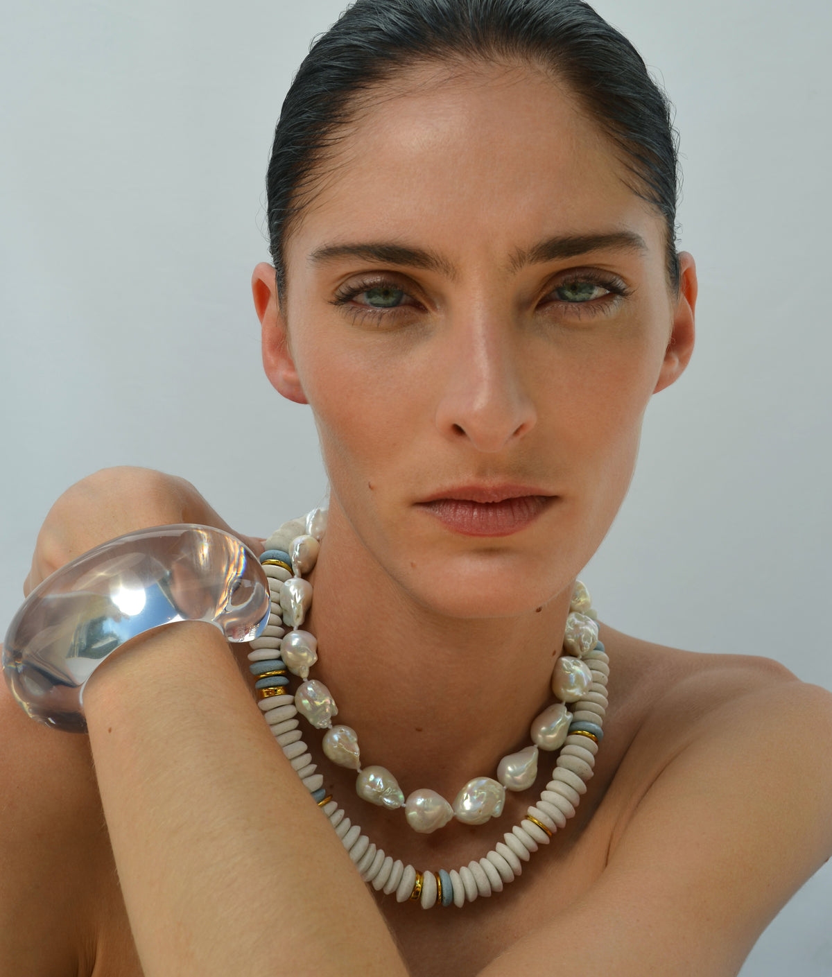 A woman with slicked-back hair wears the Laguna Necklace by Lizzie Fortunato and a large clear bracelet, posing against a plain background.