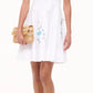 A woman wears the Staud Mini Wells Dandelion Dress, a white cotton poplin sleeveless dress with blue floral embroidery and a corset-style panel, holding a woven clutch and wearing teal sandals against a plain white background.