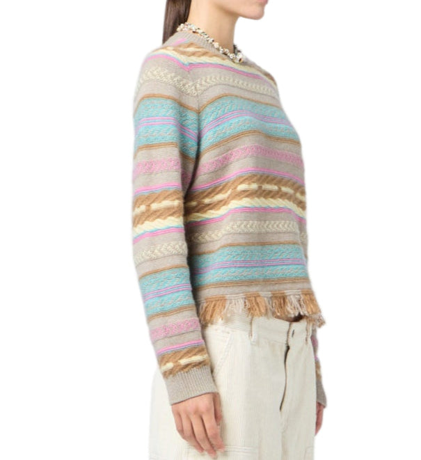 A person in the Western Stripe Crew sweater by Guest in Residence, featuring multicolored stripes and a fringe hem, paired with light-colored pants, shown from the side against a white background.