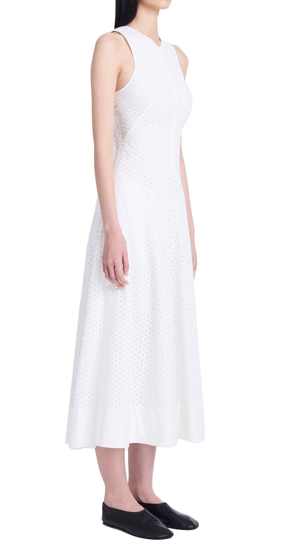 A person standing in profile, wearing a sleeveless white Proenza Schouler Juno Dress with Broderie Anglaise details and black flat shoes against a white background.