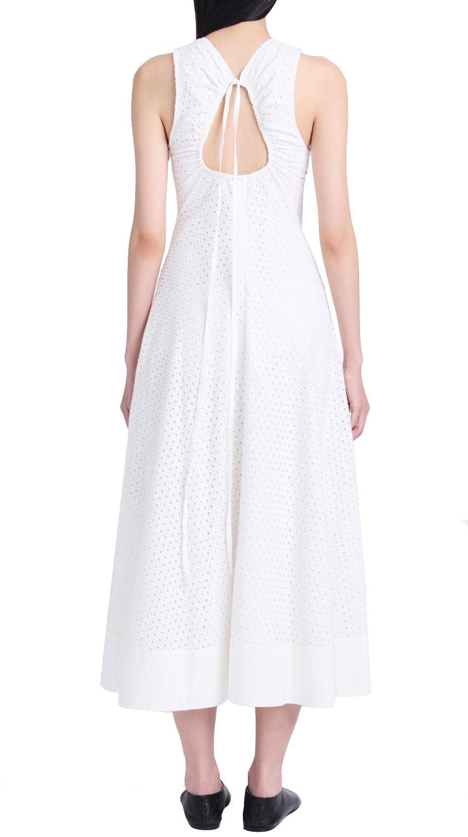 A person is wearing a sleeveless white Proenza Schouler Juno Dress with a keyhole back and a tie closure. The dress, adorned with an embroidered overlay, has a textured pattern and reaches mid-calf. They are also wearing black shoes.