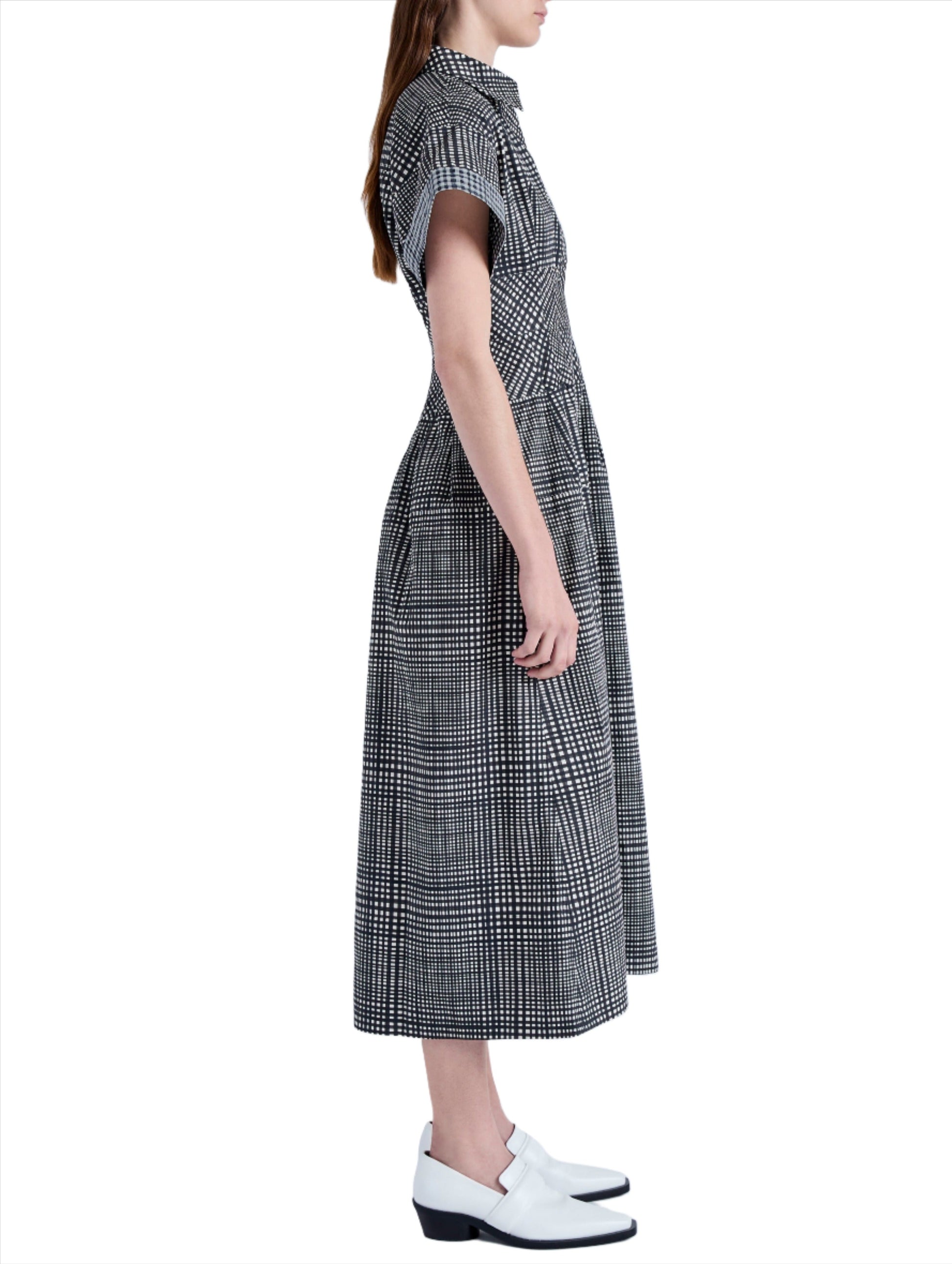 Side profile of a person wearing the Proenza Schouler Balston Grid Poplin Dress by Proenza Schouler White Label, featuring a black and white checkered A-line design with a point collar, paired with white shoes against a plain background.