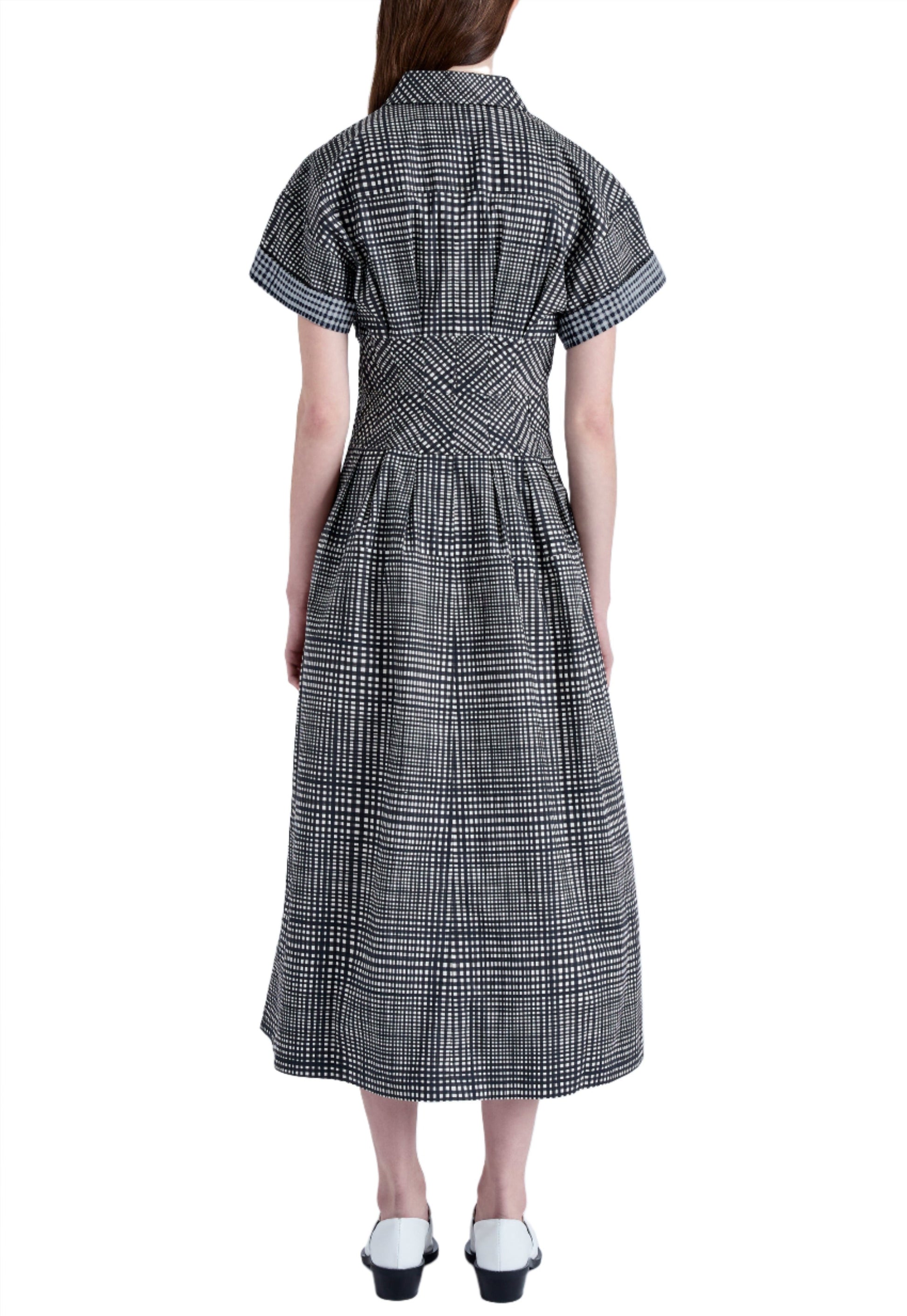 Rear view of a person wearing the Proenza Schouler Balston Grid Poplin Dress by Proenza Schouler White Label, a 100% cotton A-line dress with a black and white checkered pattern, short sleeves, and a calf-length hem, paired with matching black and white shoes.