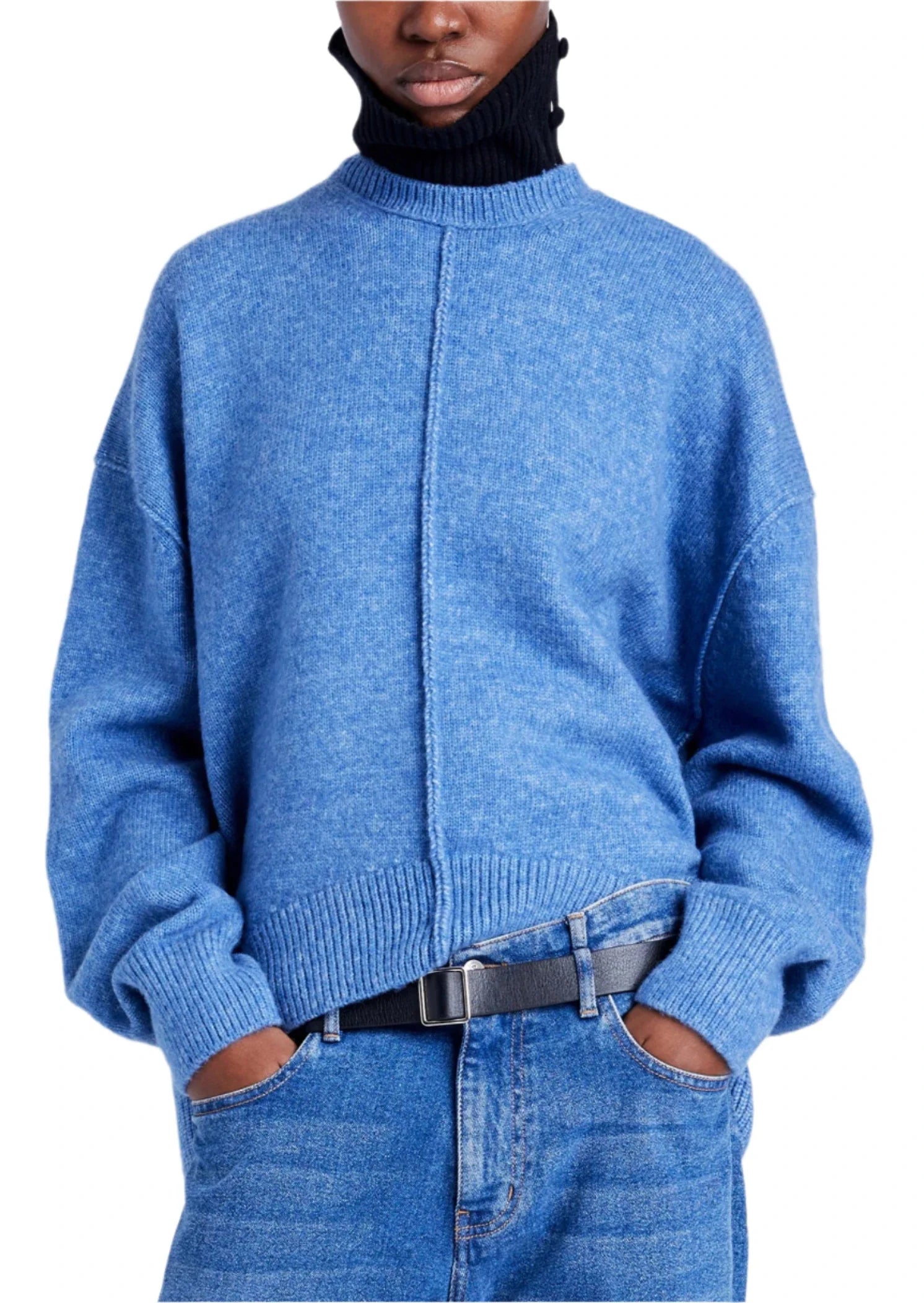 Sporting a Proenza Schouler Kellen Wool Sweater, this person embraces a relaxed fit with a blue sweater over a black turtleneck, seamlessly paired with blue jeans and a black belt. Hands tucked casually in pockets complete the effortlessly chic ensemble.