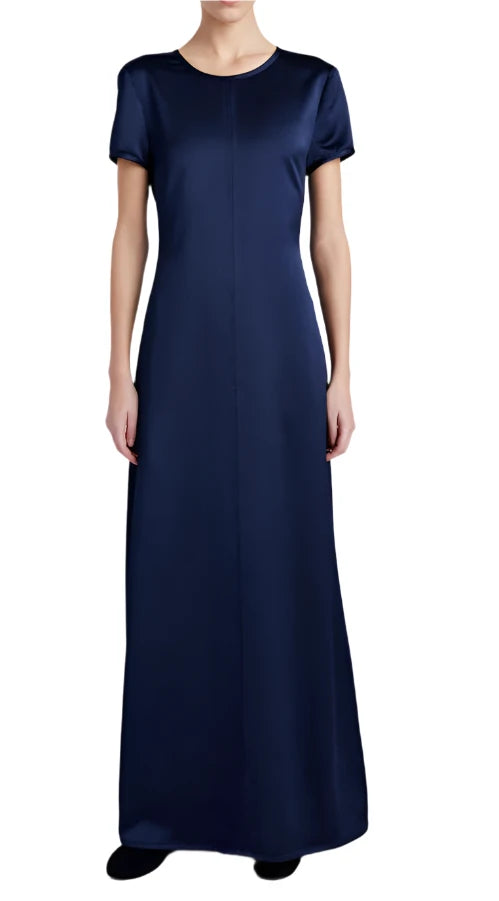 The person is dressed in a floor-length, navy blue Proenza Schouler White Label Delphine Satin Dress, which features short sleeves and an elegant A-line silhouette, standing against a plain white background.