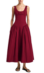 A woman in the Proenza Schouler White Label Kendall Dress—a sleeveless, ankle-length deep red dress with a scoop neckline and gathered side panels—wears black sandals and stands against a white background.