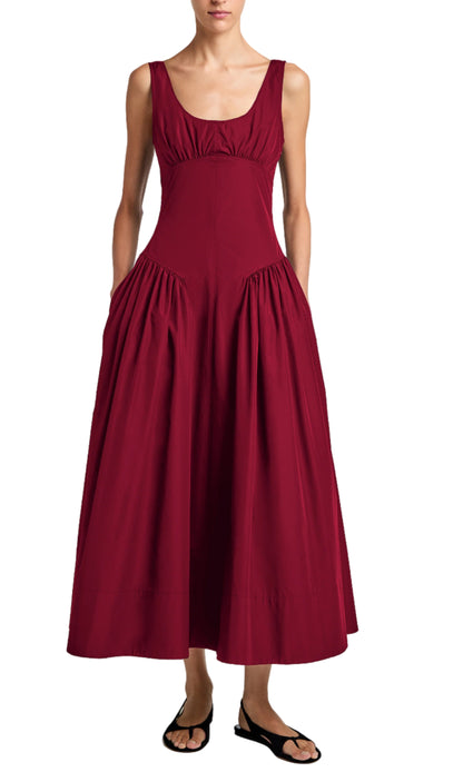 A woman in the Proenza Schouler White Label Kendall Dress—a sleeveless, ankle-length deep red dress with a scoop neckline and gathered side panels—wears black sandals and stands against a white background.